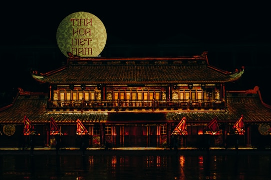 A traditional Vietnamese building illuminated at night with an intricate roof and multiple windows, adorned with red and gold lights. A large patterned circle with text in Vietnamese is displayed in the background, and several red and yellow flags are situated in front of the building.