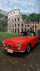 A bright red classic Alfa Romeo 2600 sports car is parked indoors on a cobblestone surface. Behind the car is a large mural depicting the Colosseum, green hills, and blue sky with scattered clouds, giving the impression of the Italian countryside.