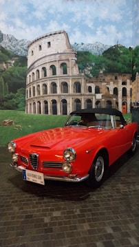 A bright red classic Alfa Romeo 2600 sports car is parked indoors on a cobblestone surface. Behind the car is a large mural depicting the Colosseum, green hills, and blue sky with scattered clouds, giving the impression of the Italian countryside.