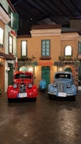 Two vintage Fiat 1100 E cars, one red and one blue, are parked in front of a quaint building with green shutters and a sign that reads 'Motel'. A blue scooter is positioned between them. The scene is reminiscent of a charming Italian street with its warm-toned facades and potted plants.