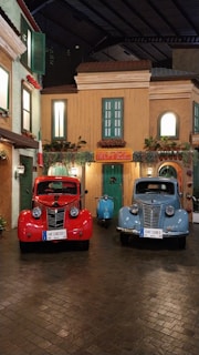 Two vintage Fiat 1100 E cars, one red and one blue, are parked in front of a quaint building with green shutters and a sign that reads 'Motel'. A blue scooter is positioned between them. The scene is reminiscent of a charming Italian street with its warm-toned facades and potted plants.
