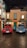 Two vintage Fiat 1100 E cars, one red and one blue, are parked in front of a quaint building with green shutters and a sign that reads 'Motel'. A blue scooter is positioned between them. The scene is reminiscent of a charming Italian street with its warm-toned facades and potted plants.