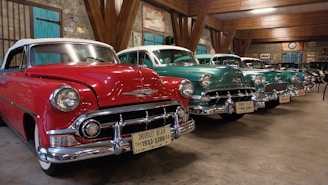 Several classic Chevrolet cars are parked indoors in a line, showcasing their vintage designs and colors. The red car in the foreground is particularly striking with chrome accents and a distinctive grill. The other cars, painted in shades of teal and green, are evenly spaced and enhance the nostalgic atmosphere of the room. The setting appears to be a well-lit garage or showroom with rustic wooden beams and various vintage automotive signs adorning the walls.