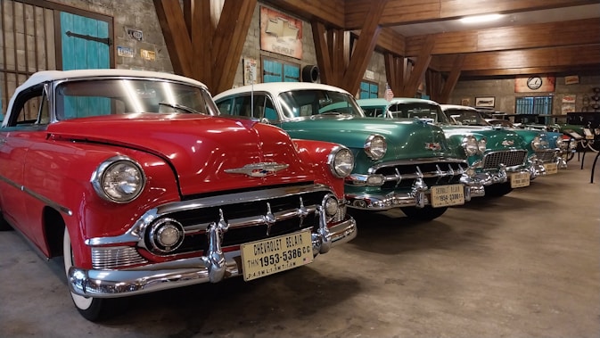 Several classic Chevrolet cars are parked indoors in a line, showcasing their vintage designs and colors. The red car in the foreground is particularly striking with chrome accents and a distinctive grill. The other cars, painted in shades of teal and green, are evenly spaced and enhance the nostalgic atmosphere of the room. The setting appears to be a well-lit garage or showroom with rustic wooden beams and various vintage automotive signs adorning the walls.