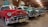Several classic Chevrolet cars are parked indoors in a line, showcasing their vintage designs and colors. The red car in the foreground is particularly striking with chrome accents and a distinctive grill. The other cars, painted in shades of teal and green, are evenly spaced and enhance the nostalgic atmosphere of the room. The setting appears to be a well-lit garage or showroom with rustic wooden beams and various vintage automotive signs adorning the walls.