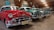 Several classic Chevrolet cars are parked indoors in a line, showcasing their vintage designs and colors. The red car in the foreground is particularly striking with chrome accents and a distinctive grill. The other cars, painted in shades of teal and green, are evenly spaced and enhance the nostalgic atmosphere of the room. The setting appears to be a well-lit garage or showroom with rustic wooden beams and various vintage automotive signs adorning the walls.