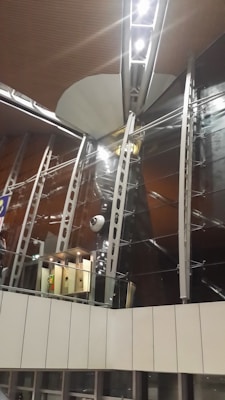 A modern interior with tall, metallic beams and large glass panels. The structure features sleek, industrial design elements, including exposed lighting and reflective surfaces. There is a section resembling an elevator area below a prominent overhang.