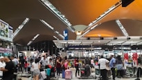 A busy airport terminal filled with travelers pushing luggage trolleys. The architectural ceiling features triangular panels with metal beams, and there are overhead lights illuminating the space. People are gathered near check-in counters, and there are signs hanging from the ceiling with letters marking various sections.