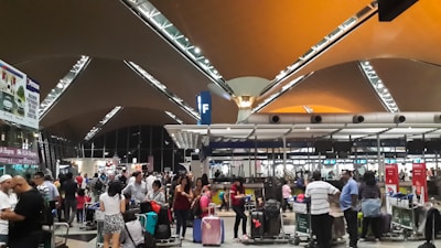 A busy airport terminal filled with travelers pushing luggage trolleys. The architectural ceiling features triangular panels with metal beams, and there are overhead lights illuminating the space. People are gathered near check-in counters, and there are signs hanging from the ceiling with letters marking various sections.