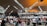 A busy airport terminal filled with travelers pushing luggage trolleys. The architectural ceiling features triangular panels with metal beams, and there are overhead lights illuminating the space. People are gathered near check-in counters, and there are signs hanging from the ceiling with letters marking various sections.