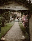 A gravel pathway lined with well-maintained grass and flower bushes leads to a hotel building in the background. An arched entrance with a sign that reads 'La Fontana Family Hotel' frames the path. Neatly trimmed trees and lampposts are present along the pathway. The atmosphere is serene and welcoming.