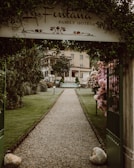 A gravel pathway lined with well-maintained grass and flower bushes leads to a hotel building in the background. An arched entrance with a sign that reads 'La Fontana Family Hotel' frames the path. Neatly trimmed trees and lampposts are present along the pathway. The atmosphere is serene and welcoming.
