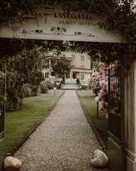 A gravel pathway lined with well-maintained grass and flower bushes leads to a hotel building in the background. An arched entrance with a sign that reads 'La Fontana Family Hotel' frames the path. Neatly trimmed trees and lampposts are present along the pathway. The atmosphere is serene and welcoming.