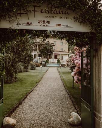 A gravel pathway lined with well-maintained grass and flower bushes leads to a hotel building in the background. An arched entrance with a sign that reads 'La Fontana Family Hotel' frames the path. Neatly trimmed trees and lampposts are present along the pathway. The atmosphere is serene and welcoming.