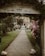 A gravel pathway lined with well-maintained grass and flower bushes leads to a hotel building in the background. An arched entrance with a sign that reads 'La Fontana Family Hotel' frames the path. Neatly trimmed trees and lampposts are present along the pathway. The atmosphere is serene and welcoming.