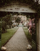 A gravel pathway lined with well-maintained grass and flower bushes leads to a hotel building in the background. An arched entrance with a sign that reads 'La Fontana Family Hotel' frames the path. Neatly trimmed trees and lampposts are present along the pathway. The atmosphere is serene and welcoming.