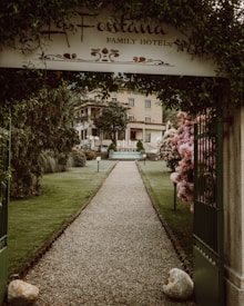A gravel pathway lined with well-maintained grass and flower bushes leads to a hotel building in the background. An arched entrance with a sign that reads 'La Fontana Family Hotel' frames the path. Neatly trimmed trees and lampposts are present along the pathway. The atmosphere is serene and welcoming.