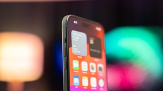 A close-up view of a smartphone showing the home screen filled with app icons and widgets. The screen displays clear details such as weather information and icons for apps like FaceTime and Mail. The background is blurred with vibrant colors, adding a modern and dynamic feel.