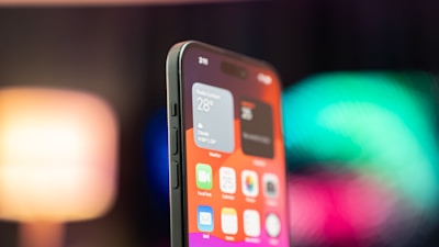 A close-up view of a smartphone showing the home screen filled with app icons and widgets. The screen displays clear details such as weather information and icons for apps like FaceTime and Mail. The background is blurred with vibrant colors, adding a modern and dynamic feel.