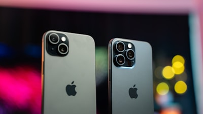 Two modern smartphones with sleek designs are positioned side by side against a blurred, colorful background featuring bokeh lights. Both phones have multiple camera lenses on the back and a metallic finish, with the iconic Apple logo displayed prominently.