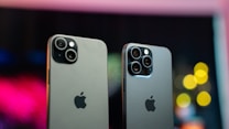 Two modern smartphones with sleek designs are positioned side by side against a blurred, colorful background featuring bokeh lights. Both phones have multiple camera lenses on the back and a metallic finish, with the iconic Apple logo displayed prominently.