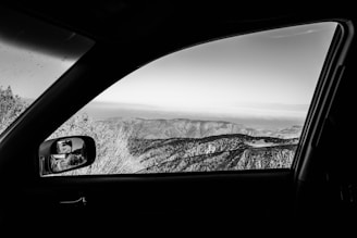 A black and white photograph taken from inside a car, focusing on the landscape beyond the vehicle's side window. The scene outside includes rugged hills and sparse vegetation under a clear sky. The car's interior, mirror, and window frame the view, adding depth to the composition.