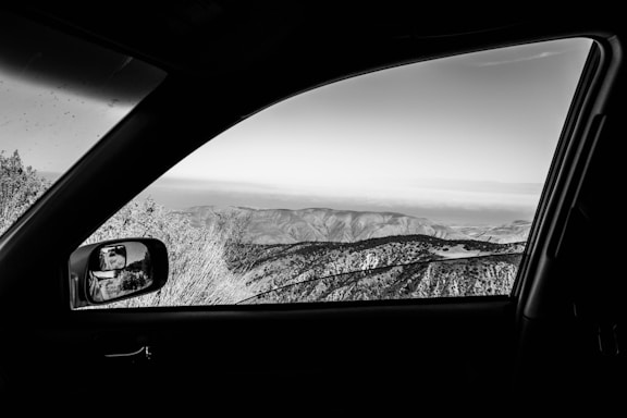 A black and white photograph taken from inside a car, focusing on the landscape beyond the vehicle's side window. The scene outside includes rugged hills and sparse vegetation under a clear sky. The car's interior, mirror, and window frame the view, adding depth to the composition.