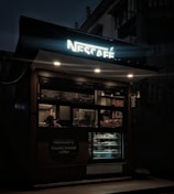 A small, illuminated coffee kiosk with a glowing Nescafe sign at night. The kiosk has a display of pastries and a menu visible through the glass. A person is seen inside the kiosk preparing or serving drinks.