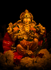 A vividly colored statue of the Hindu deity Ganesha, featuring intricate adornments and surrounded by flowers. The figure is illuminated with warm lighting, highlighting the golden crown, expressive eyes, and detailed ornaments. The floral garlands and offerings add to the rich, festive atmosphere.