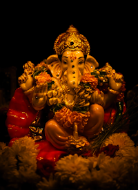 A vividly colored statue of the Hindu deity Ganesha, featuring intricate adornments and surrounded by flowers. The figure is illuminated with warm lighting, highlighting the golden crown, expressive eyes, and detailed ornaments. The floral garlands and offerings add to the rich, festive atmosphere.