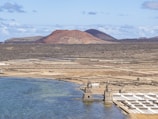 An expansive, arid landscape featuring geometrically arranged salt flats adjacent to a body of water. Stone structures with towers are situated near the shoreline, and the background includes rugged terrain with reddish-brown volcanic hills under a partly cloudy sky.