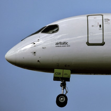 A close-up view of the front section of an aircraft featuring the logo and text 'airBaltic' and 'AIRBUS A220-300' on its body. The landing gear is extended, and the scene is set against a clear blue sky.