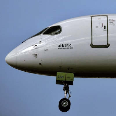 A close-up view of the front section of an aircraft featuring the logo and text 'airBaltic' and 'AIRBUS A220-300' on its body. The landing gear is extended, and the scene is set against a clear blue sky.
