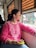 A person with dark hair tied back is sitting by a window in a vehicle, looking outside with a contemplative expression. They are wearing a vibrant pink kurta with intricate white embroidery and holding a bright green smartphone in their hand. The window has metal grilles, and there are blurred urban scenes visible outside.