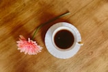 A serene coffee cup beside a pink flower on a white tabletop.