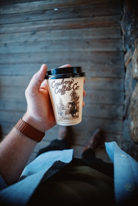 A hand holding a cup of Cowboy Coffee Co. against a wooden background. The person is wearing a watch with a brown strap and a light blue shirt.