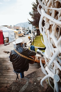 A person wearing a leather jacket and a cowboy hat is playing a guitar on a wooden deck. The setting includes a rustic street with tents, and white antlers are prominently displayed in the foreground.