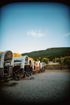A series of covered wagons are lined up on a gravel area, surrounded by wooden fences and set against a backdrop of lush green hills and a clear blue sky. A person is seated on the steps of one of the wagons, wearing a cowboy hat and casual attire. The setting appears to be rustic and peaceful, suggesting a historical or Western theme.