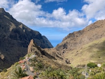 A scenic landscape of a mountainous valley with towering cliffs on either side. A winding road lined with small houses and palm trees leads through the valley, with a view of the ocean in the distance. Clear blue skies with scattered white clouds enhance the natural beauty of the setting.