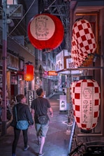 A narrow, dimly lit street in what appears to be a Japanese city is adorned with large, decorative lanterns hanging overhead. The lanterns cast a warm glow over the scene, displaying Japanese characters. A couple is walking side by side, adding a sense of movement and life to the quiet alley. The storefronts are lined with signs and posters, suggesting a lively neighborhood.