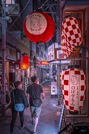 A narrow, dimly lit street in what appears to be a Japanese city is adorned with large, decorative lanterns hanging overhead. The lanterns cast a warm glow over the scene, displaying Japanese characters. A couple is walking side by side, adding a sense of movement and life to the quiet alley. The storefronts are lined with signs and posters, suggesting a lively neighborhood.