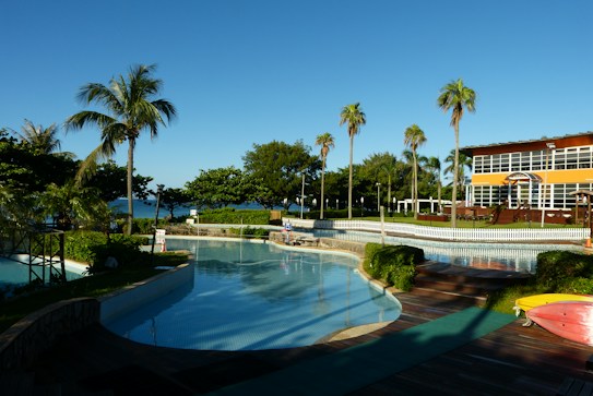 A serene outdoor scene featuring a swimming pool surrounded by lush greenery and palm trees. In the background, there is a multi-story building with large windows and a yellow facade. The blue sky is clear, and the ocean can be seen in the distance. Wooden decks and pathways add a warm tone to the landscape.