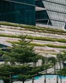 A modern building with a sloped green roof featuring multiple levels of vegetation. Large glass windows are visible in the background, and there are several trees and landscaped greenery at the base.
