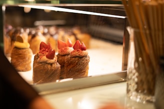 A display case in a bakery showcasing a variety of pastries topped with cream and fresh strawberries. The desserts are elegantly arranged under warm lighting, enhancing their golden brown hues and inviting presentation. The background is softly blurred, focusing attention on the intricate details of the pastries.