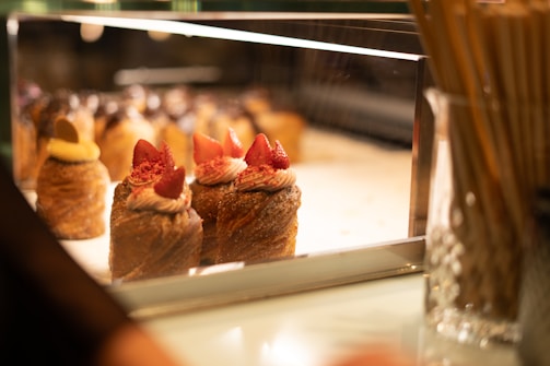 A display case in a bakery showcasing a variety of pastries topped with cream and fresh strawberries. The desserts are elegantly arranged under warm lighting, enhancing their golden brown hues and inviting presentation. The background is softly blurred, focusing attention on the intricate details of the pastries.