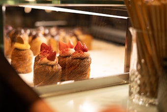 A display case in a bakery showcasing a variety of pastries topped with cream and fresh strawberries. The desserts are elegantly arranged under warm lighting, enhancing their golden brown hues and inviting presentation. The background is softly blurred, focusing attention on the intricate details of the pastries.