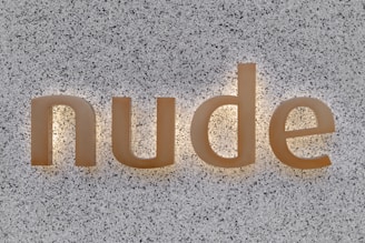 The word 'nude' is displayed in lowercase letters on a speckled gray background. The letters appear to be made of a light-colored material and are illuminated from the back, creating a soft glow around them.