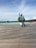 A person is walking on a wooden boardwalk by the water, with a row of overwater bungalows in the background. The person is dressed casually in jeans, a green shirt, and a checkered shirt, and appears to be posing with their hand raised. There are some planters with greenery along the boardwalk.