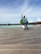 A person is walking on a wooden boardwalk by the water, with a row of overwater bungalows in the background. The person is dressed casually in jeans, a green shirt, and a checkered shirt, and appears to be posing with their hand raised. There are some planters with greenery along the boardwalk.