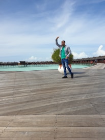 A person is walking on a wooden boardwalk by the water, with a row of overwater bungalows in the background. The person is dressed casually in jeans, a green shirt, and a checkered shirt, and appears to be posing with their hand raised. There are some planters with greenery along the boardwalk.